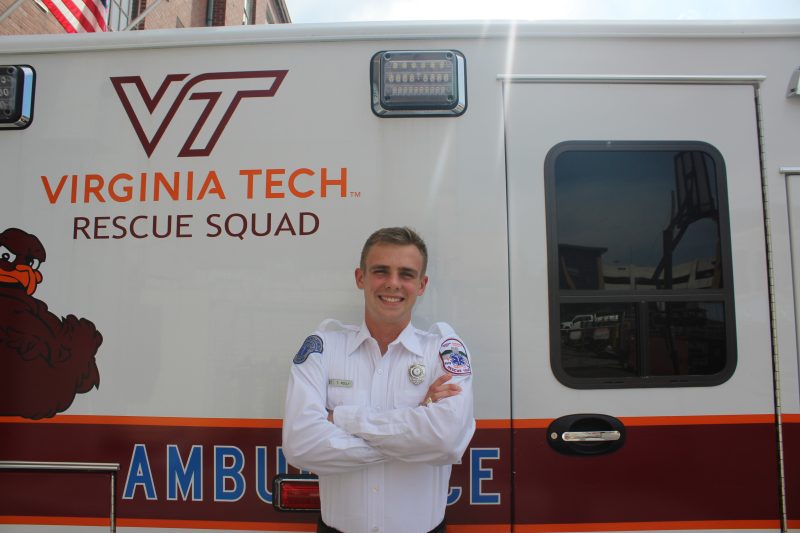 Thomas Feely smiles in a white uniform shirt stands with arms crossed in front of an ambulance featuring the Virginia Tech Rescue Squad logo and a HokieBird graphic.