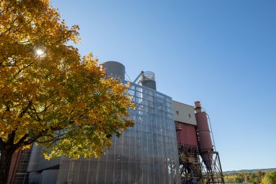 Grey grating surrounds the red brick and red painted metal Virginia Tech Power Plant. It's surrounded by trees just beginning to turn yellow from fall weather on a sunny day.
